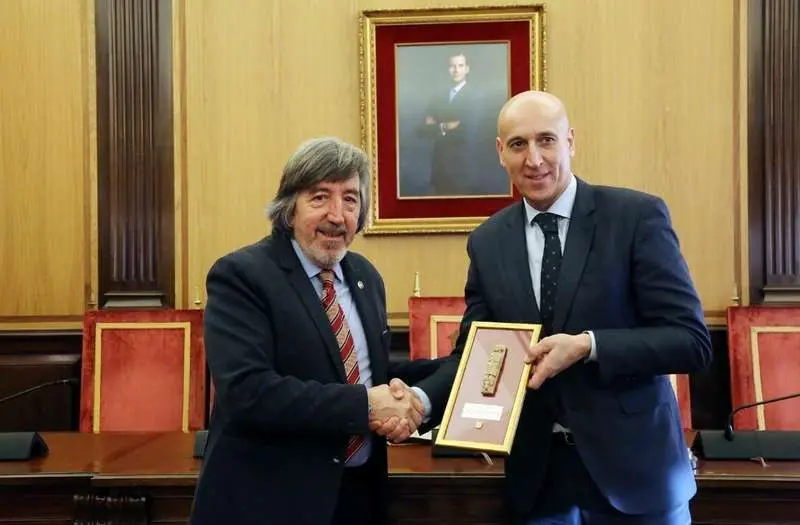 Dos hombres con traje estrechándose la mano y sosteniendo una placa conmemorativa en un despacho oficial